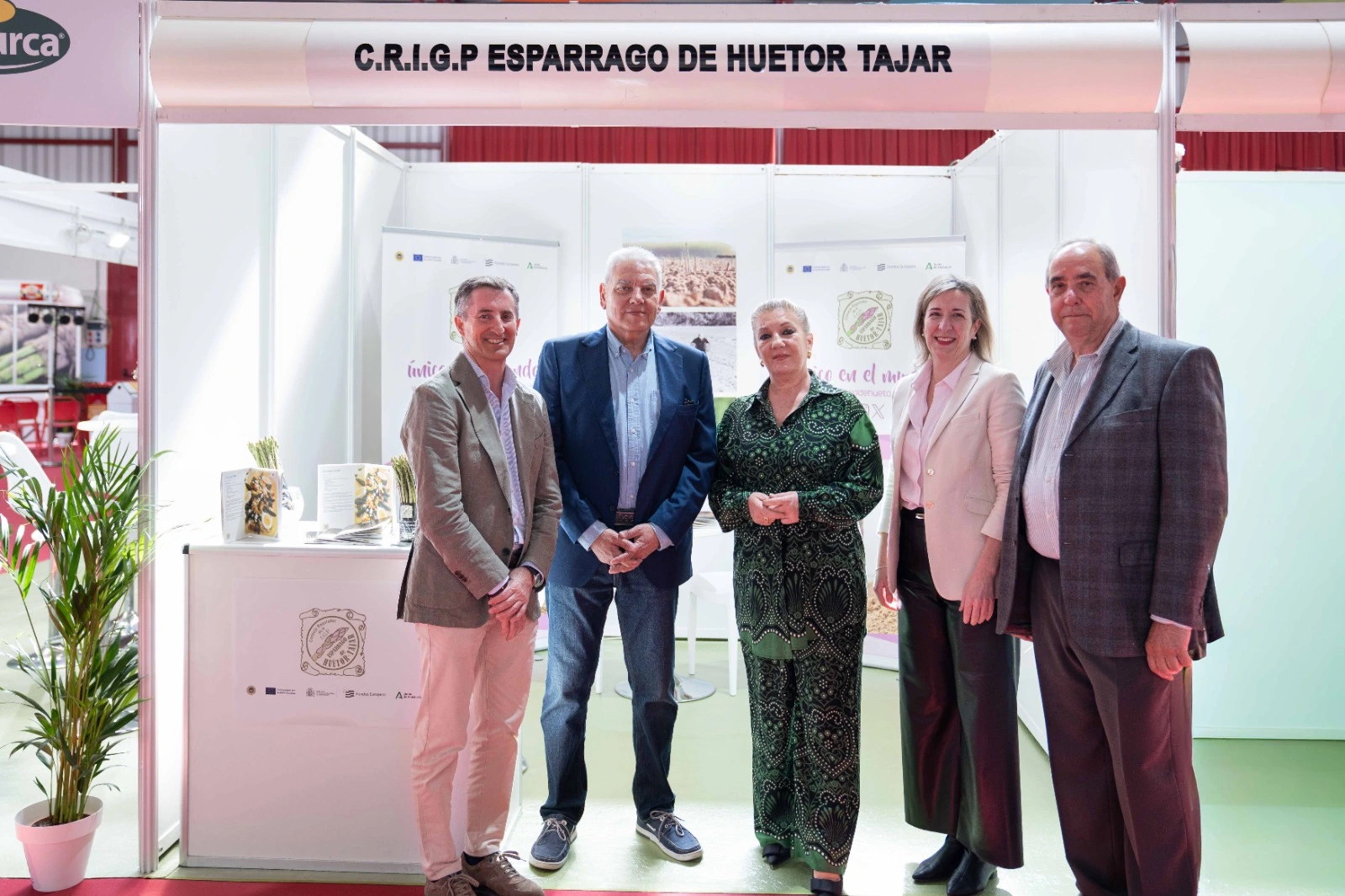 Grupo de autoridades, representantes del sector y la mascota con forma de espárrago posando en el stand de la IGP Espárrago de Huétor Tájar durante la III Feria Gastronómica.